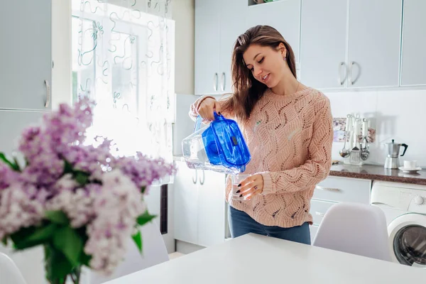 Mujer vertiendo agua filtrada de la jarra de filtro en vidrio en la cocina. Diseño moderno de la cocina. Estilo de vida saludable — Foto de Stock
