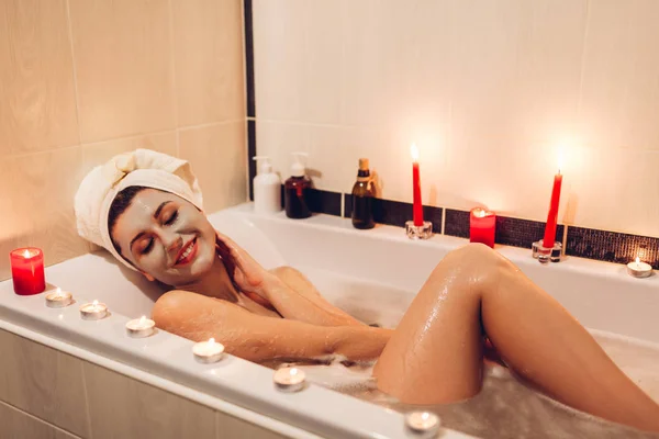 Happy young woman relaxing in bath tub with foam and candles with facial clay mask applied at home. Sexy lady chilling in hotel bathroom