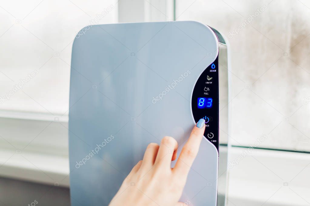Woman turns dehumidifier on using touch panel by wet window. Humidity indicator shows high level of dampness in flat. Air dryer