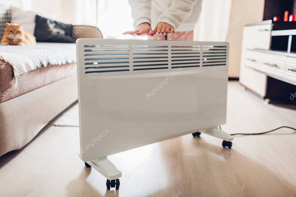 Using heater at home in winter. Woman warming her hands by device and wearing warm clothes. Heating season.