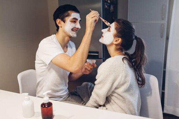 Man applying clay mask on his girlfriend's face. Young loving couple taking care of skin on kitchen at home