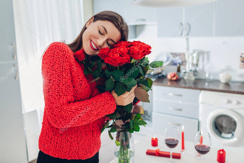 Young woman found bouquet of roses, wine and gift box on kitchen. Happy girl hugging flowers. Valentines day surprise