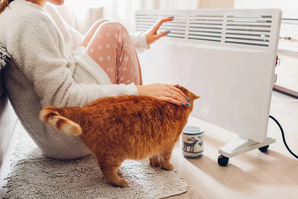 Uso del riscaldatore a casa in inverno. Donna che si scalda le mani con il gatto. Stagione di riscaldamento . — Foto Stock