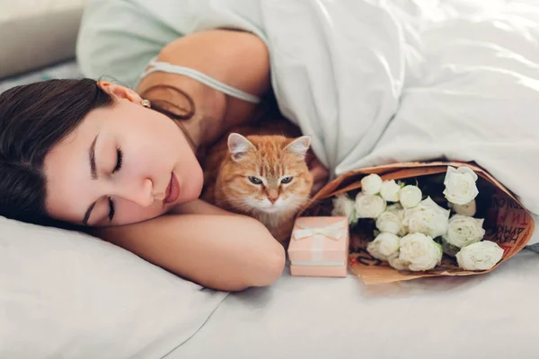 Young woman sleeping near bouquet of roses and gift box in bed with cat. Present left by boyfriend for Women's day