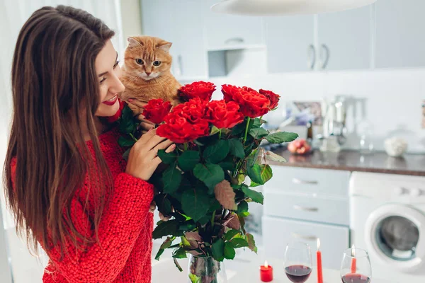 Ung kvinna hittade röda rosor med ljus, vin och gåva rutan på kök. Glad tjej att lukta blommor med katt. — Stockfoto