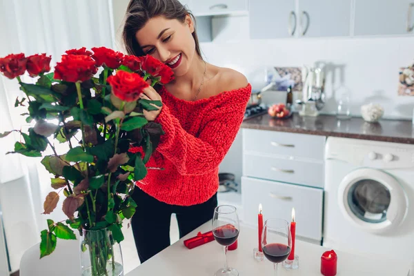 Молода жінка знайшла червоні троянди зі свічкою, вином і подарунковою коробкою на кухні. Щаслива дівчина пахне квітами. Жіночий день — стокове фото