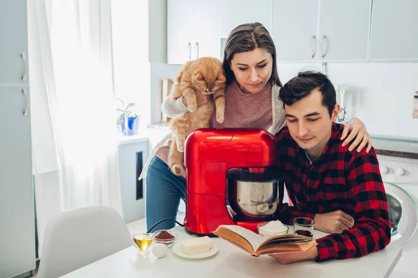 Jonge gelukkige paar koken op keuken met keukenmachine bedrijf kat. Vrouw en man lezen recept en knuffelen — Stockfoto