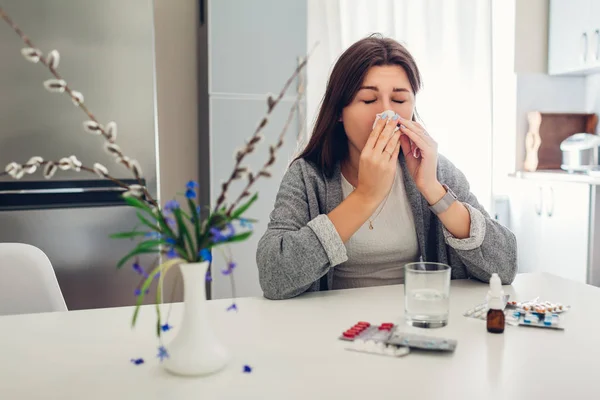 Lente allergie. Jonge vrouw niezen vanwege bloemen omgeven met pillen op keuken. Seizoensgebonden allergie concept. — Stockfoto