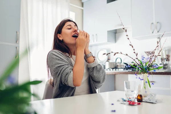 Alergia de primavera. Mujer joven estornudando debido a las flores rodeadas de pastillas en la cocina. Concepto de alergia estacional . — Foto de Stock