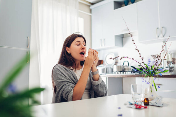 Spring allergy. Young woman sneezing because of flowers surrounded with pills on kitchen. Seasonal allergy concept.