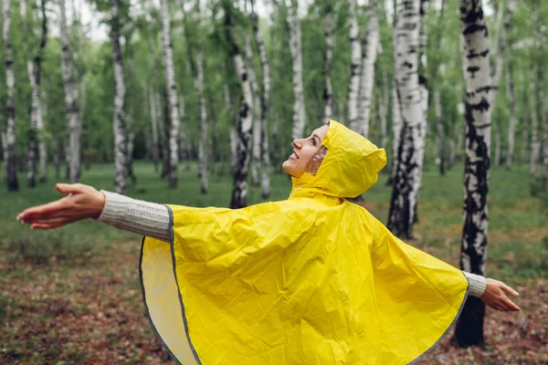 Happy young woman in yellow raincoat walking in spring forest under rain and having fun raising arms