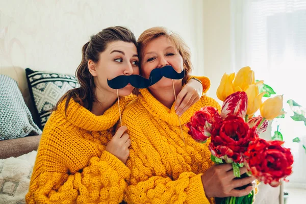 Mère aînée et sa fille adulte prenant selfie avec des fleurs en utilisant des accessoires de photomaton à la maison. Concept de fête des mères . — Photo