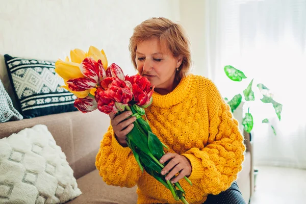 Mulher sênior cheirando buquê de tulipas de seus filhos em casa. Presente de dia de mãe — Fotografia de Stock