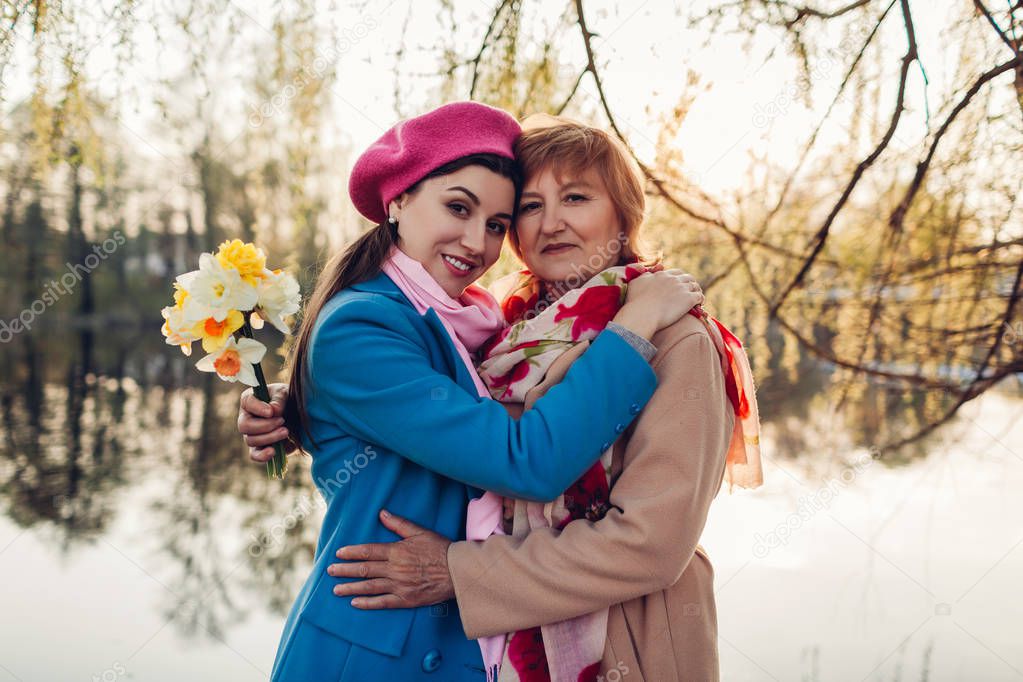 Senior mother with flowers and her adult daughter hugging by spring river. Mother's day concept. Family values