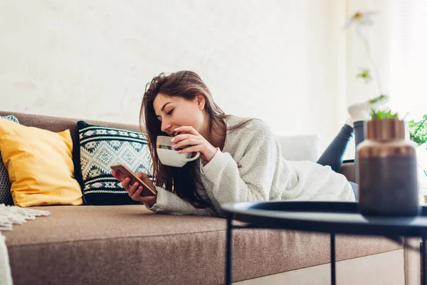 Fiatal nő pihen a nappaliban okostelefonnal és kávéval. Belső dekoráció — Stock Fotó