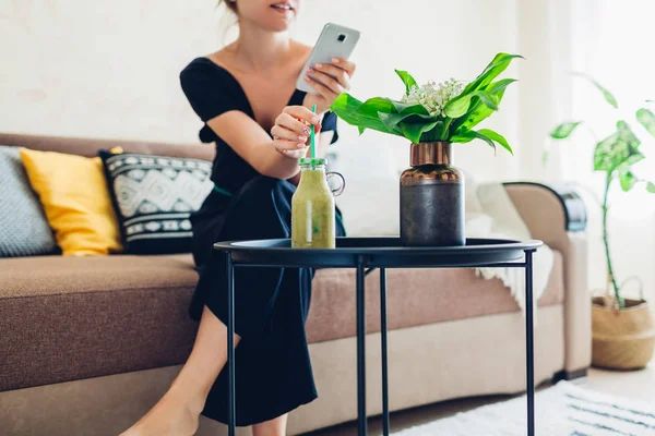 Jonge vrouw ontspannen in de woonkamer met behulp van smartphone en het drinken van smoothie. Thuis comfort en gezelligheid — Stockfoto