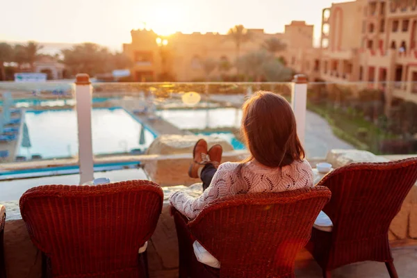 Woman relaxing at hotel balcony enjoying sunrise with swimming pool and sea view.