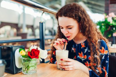 Young woman drinks coffee using straw in shopping center cafe. Girl enjoying latte and relaxing