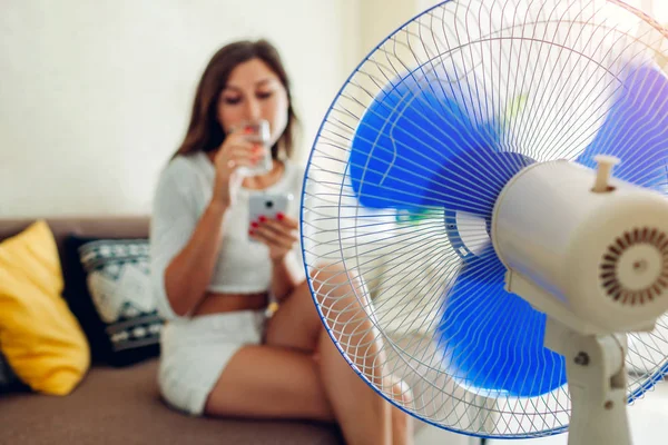 Fiatal nő lehűlés a ventilátor otthon, miközben iszik vizet, és lóg a telefon. Nyári hőség. — Stock Fotó