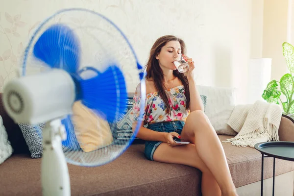 Fiatal nő lehűlés a ventilátor otthon, miközben iszik vizet, és lóg a telefon. Nyári hőség. — Stock Fotó