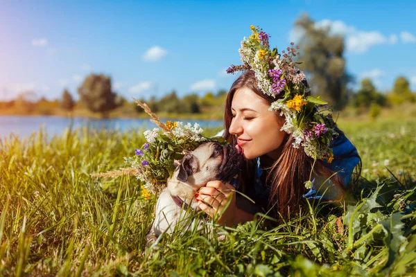 Pug dog y su amo escalofriante por el río de verano vistiendo coronas de flores. Feliz cachorro lamiendo mujer — Foto de Stock
