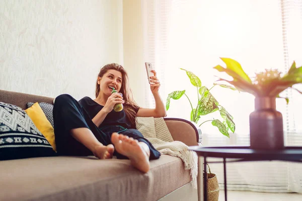 Fiatal nő pihentető nappali és ivás smoothie segítségével okostelefon. Egészséges táplálkozás — Stock Fotó