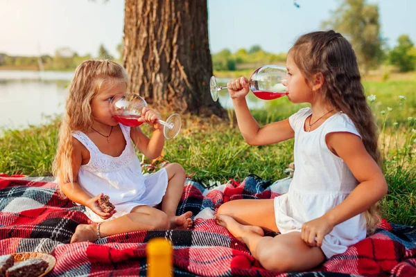 Hermanas bebiendo jugo mientras hacen picnic en el río de verano al atardecer. Niños comiendo pastel y relajándose con la familia —  Fotos de Stock
