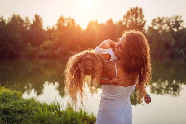 Mãe brincando e se divertindo com a filha pelo rio de verão ao pôr do sol. Mulher segurando criança e rindo. Família — Fotografia de Stock