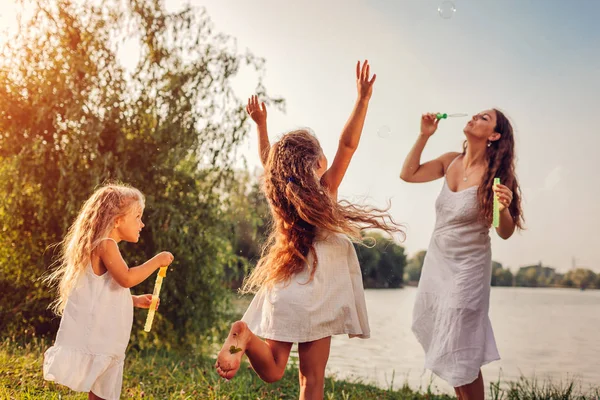 Madre ayuda a las hijas a soplar burbujas en el parque de verano. Niños divirtiéndose jugando y atrapando burbujas al aire libre —  Fotos de Stock