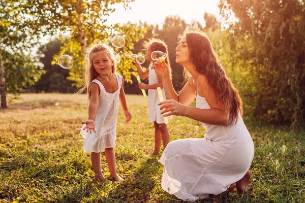 A mãe ajuda filhas a soprar bolhas no parque de verão. Crianças se divertindo brincando e pegando bolhas ao ar livre — Fotografia de Stock