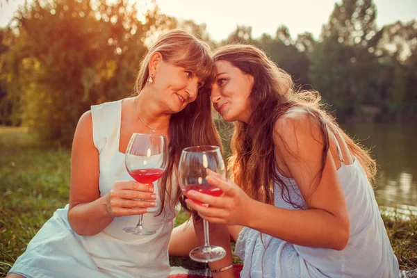 Mother drinking wine with her adult daughter by summer river at sunset. Family having picnic. Mother\'s day