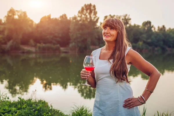 Зріла жінка насолоджується келихом вина біля річки в осінньому парку. Старша жінка захоплюється пейзажем — стокове фото