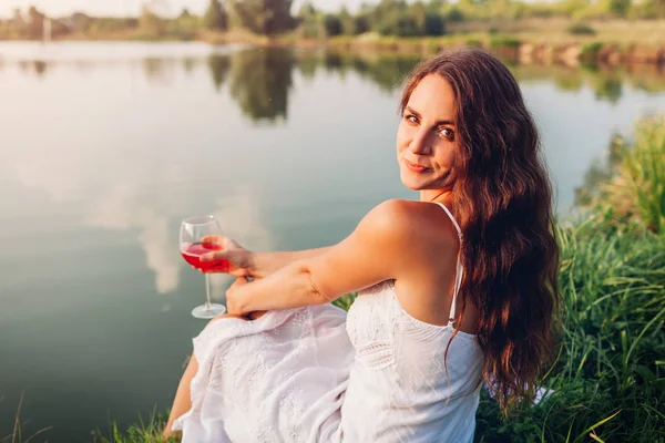 Mladá žena požívající při západu slunce sklenku vína na břehu řeky. Žena obdivující krajinu při pití — Stock fotografie