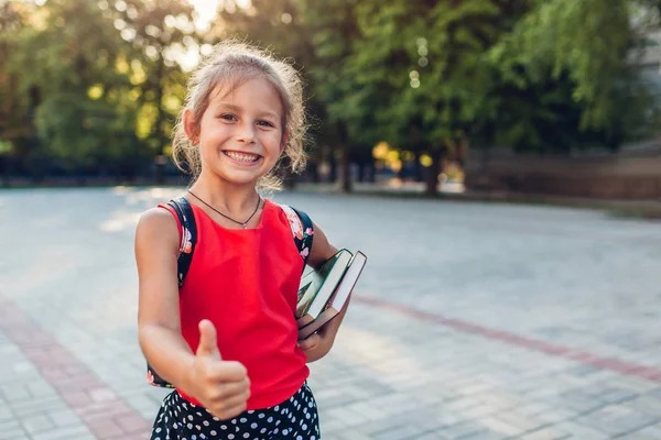 Joyeux élève portant un sac à dos et tenant des livres. Un gamin qui montre son pouce à l'école primaire. Éducation — Photo