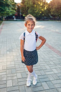 Sırt çantası ve okul üniforması giyen mutlu öğrenci kız. Çocuk kamera açık havada ilköğretim ayakta bakıyor.