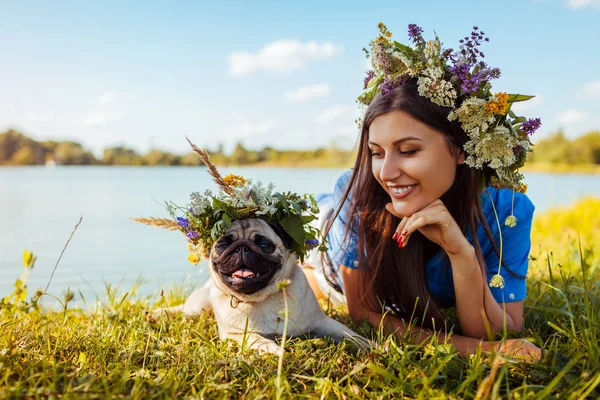 Pug dog y su amo se enfrían junto al río llevando coronas de flores. Feliz cachorro y mujer disfrutando de la naturaleza de verano al aire libre — Foto de Stock