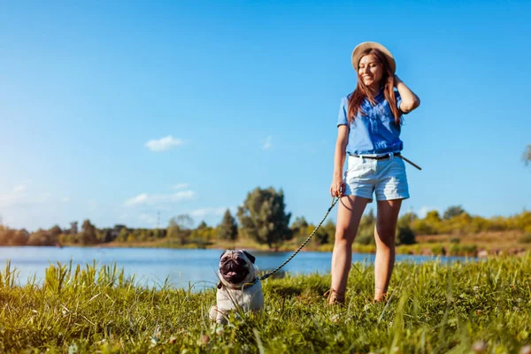 Nehir kıyısında pug köpeğiyle yürüyen mutlu bir kadın. Köpek ve kadın açık havada ürpertici