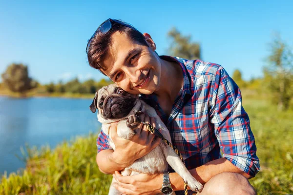 Un uomo che abbraccia un cane carlino nel parco. Uomo che cammina con animale domestico dal lago estivo — Foto Stock