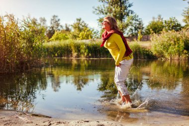 Sonbahar günü nehir kıyısında atlayan orta yaşlı bir kadın. Mutlu yaşlı bayan ormanda yürürken eğlenmek