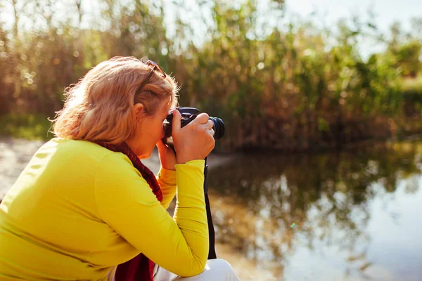 Жінка середнього віку перевіряє зображення на камерування на березі осінньої річки. Старша жінка насолоджується хобі — стокове фото
