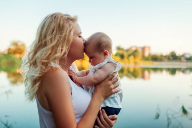 Genç anne, yaz nehrinde kız bebeği tutup öpüyor. Gün batımında çocukla yürüyen kadın