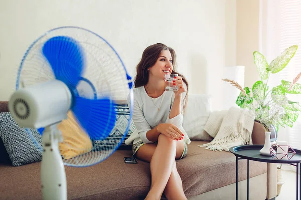 Fiatal nő lehűlés a ventilátor otthon, miközben iszik vizet, és lóg a telefon. Nyári hőség. — Stock Fotó