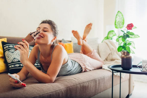 Young woman eating chocolate ice-cream in cone lying on couch at home and using smartphone. — Stock Fotó