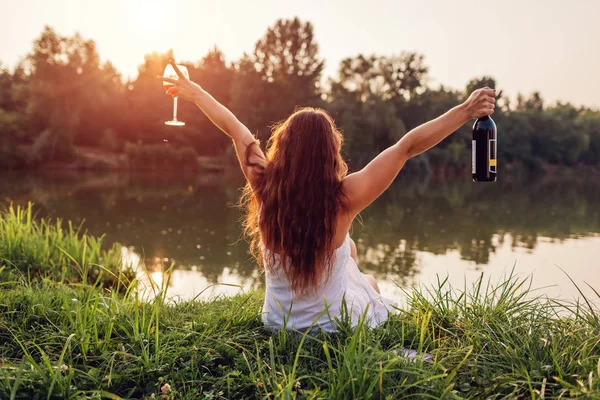Escalofriante con vino. Mujer disfrutando de una copa de vino en la orilla del río al atardecer levantando brazos y sintiéndose libre y feliz . —  Fotos de Stock
