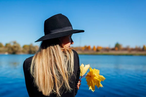 Φθινοπωρινή αύρα. Νεαρή γυναίκα χαλαρώνει δίπλα στη λίμνη κρατώντας κίτρινα φύλλα σφενδάμου — Φωτογραφία Αρχείου