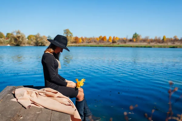 Φθινοπωρινό τοπίο Λυπηρή γυναίκα που κάθεται στην προβλήτα του ποταμού κρατώντας κίτρινα φύλλα — Φωτογραφία Αρχείου