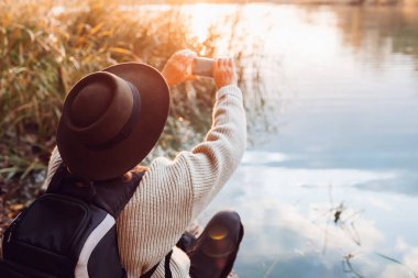 Sırt çantalı turist gün batımında akıllı telefon kullanarak fotoğraf çekiyor. Kadın sonbahar doğasına hayran bir şekilde seyahat ediyor.