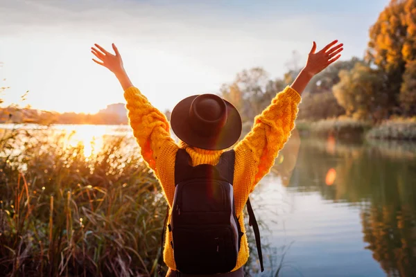 Utazó hátizsákkal pihentető őszi folyó naplementekor. Fiatal nő felemelt karok érzés szabad és boldog — Stock Fotó