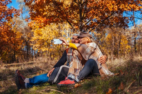 Starší pár bere selfie v podzimním parku. Šťastný muž a žena těší přírodu a objímání — Stock fotografie