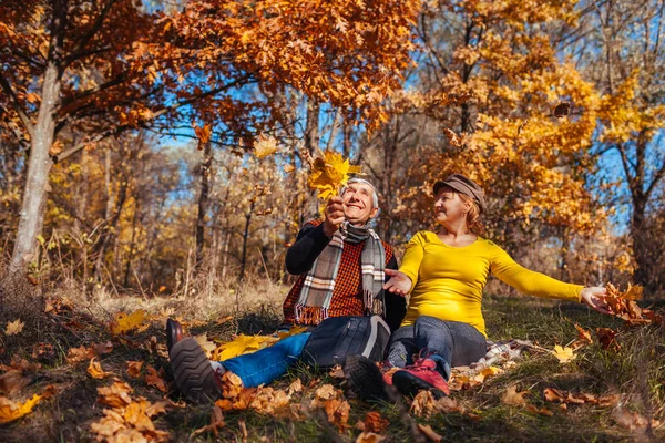 Веселье осеннего сезона. Старшая пара бросает листья сидя в парке. Мужчина и женщина отдыхают на открытом воздухе — стоковое фото